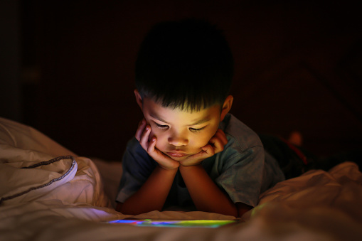 Little asian kid alone watching tablet device, lying on white duvet bed with chin on hands, in background darkness bedroom night time. Colourful bright light from screen reflex on the boy face.
