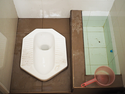 Top view Thai traditional public white ceramic squatty pot toilet with wet brown tile floor and water basin, pink plastic bucket. shiny white wall