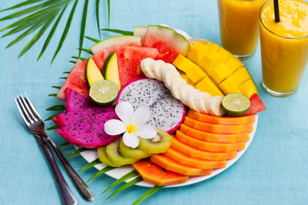 istock Tropical fruits assortment on a white plate with palm tree leaf with mango smoothie, textile blue background. Top view. Copy space 925176470