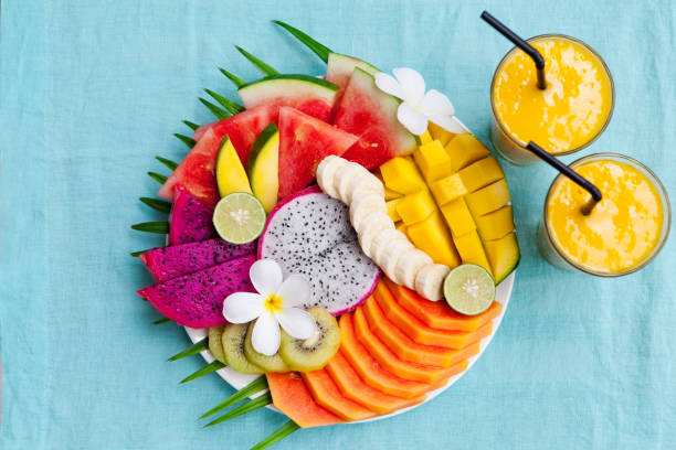 istock Tropical fruits assortment on a white plate with palm tree leaf with mango smoothie, textile blue background. Top view. Copy space 925180040