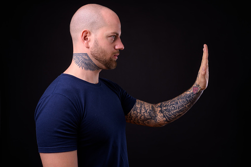 Studio shot of young handsome bearded bald man with tattoos against black background