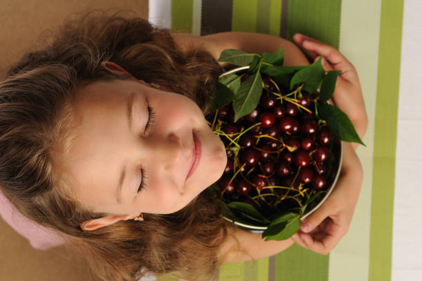 Little girl with a cherry . A happy and cheerful girl holds a pot of cherries in her hands. She is very cheerful that now she will eat her favorite berries cherries. Sweet cherry large red ripe with green leaves. The girl has big beautiful eyes.