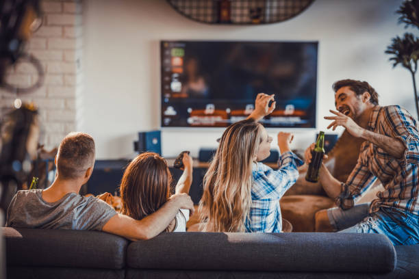 Group of happy people spending their time in the living room. Happy roommates having fun while spending their time together in the living room. Two of them are watching TV while other two are dancing. couch back stock pictures, royalty-free photos & images