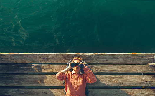 Young Beautiful Girl Lies On A Pier Near The Sea And Looks Through Binoculars On Tje Sky. Travel Search Journey Concept
