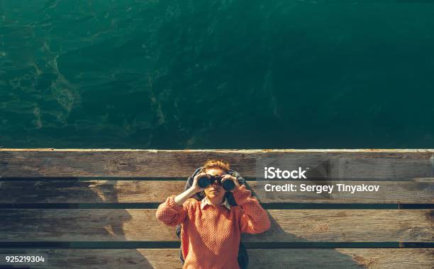 Young Beautiful Girl Lies On A Pier Near The Sea And Looks Through Binoculars On Tje Sky Travel Search Journey Concept Stock Photo - Download Image Now