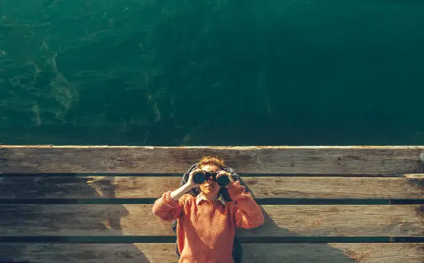 Photo of Young Beautiful Girl Lies On A Pier Near The Sea And Looks Through Binoculars On Tje Sky. Travel Search Journey Concept