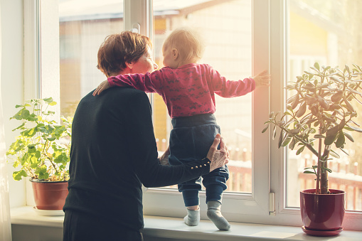 grandmother playing and taking care of child at home. standing on window sill