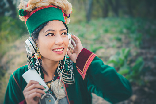 Asian, Indian, happy tribal young woman of Kinnaur, Himachal Pradesh, India in her village. She is holding smart phone, listening music or podcast through headphones and enjoying fresh air in rural area at day time. She is in their traditional formal dress and jewelry.