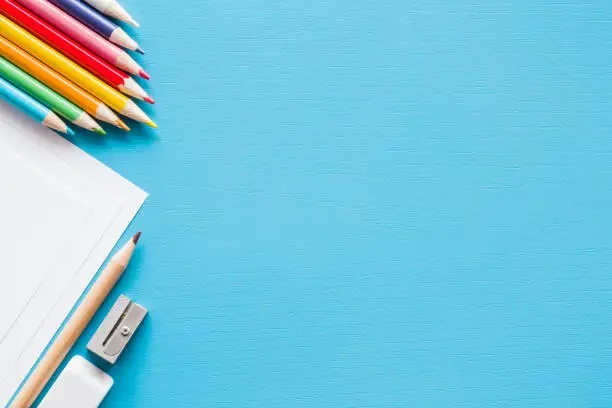 Colorful pencils, white papers and metal pencil sharpener. Empty place for text or drawing on the blue background. Childhood creative art concept. Flat lay.