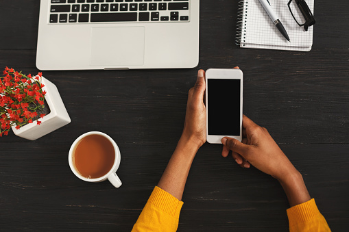 Black female hands holding smartphone with blank screen for advertisement. Top view of african-american hands, laptop keyboard, coffee, mobile on dark wooden table background, copy space