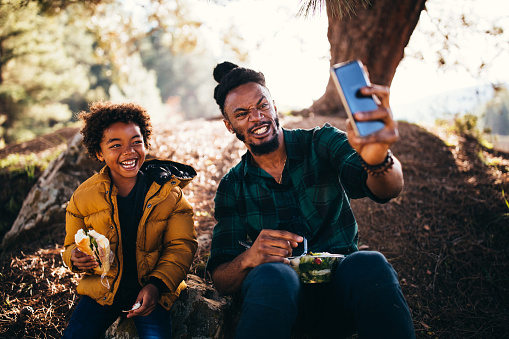 Happy father and son having fun eating lunch in forest and taking selfies on smartphone