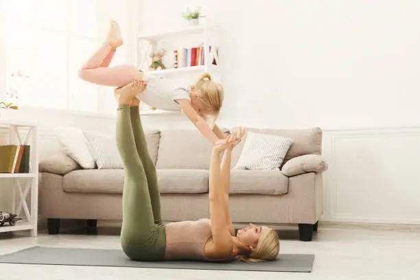 Young woman and little girl doing partner yoga flying pose, exercising together at home. Mother and daughter activity, acroyoga. Trust and support concept