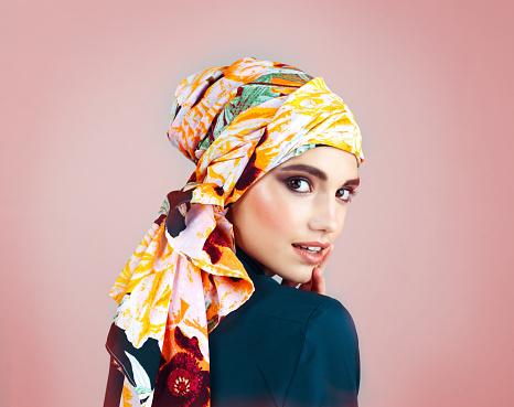 Studio portrait of a confident young woman wearing a colorful head scarf while posing against a pink background