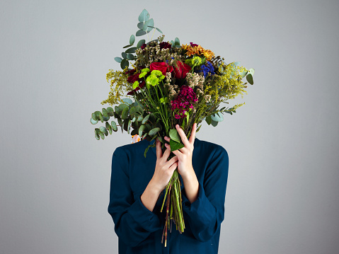 Studio shot of an unrecognizable woman holding a bouquet of flowers in front of her face while standing against a grey background