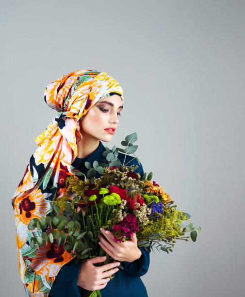 Studio shot of a confident young woman wearing a colorful head scarf and holding a bouquet of flowers against a grey background