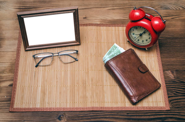 Empty photo frame with copy space, red alarm clock and wallet with money on wooden table background.
