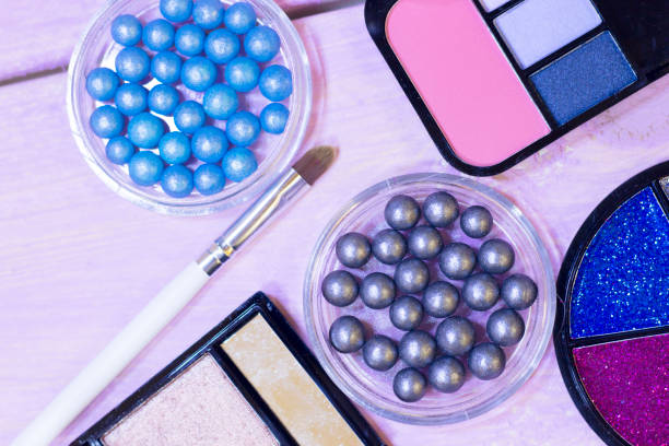 Grey and blue pearl eyeshadow in balls, eyeshadow palettes, a box of colorful glitter and brushes on white wooden background, top view, close up