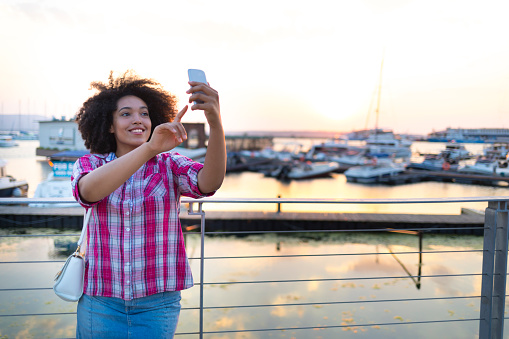 African woman taking selfie on yacht harbor, at sunset.