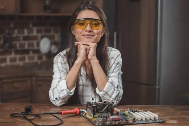 Young woman in protective glasses sitting at table with motherboard on it