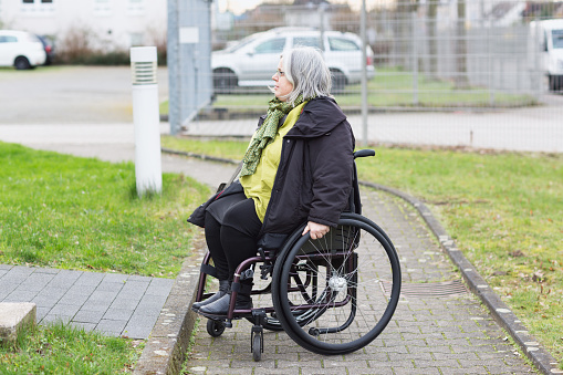woman in wheelchair im front of a step, easy fo a walker, difficult to get over when sitting in wheelchiar. Urban surrounding, parking lot in background