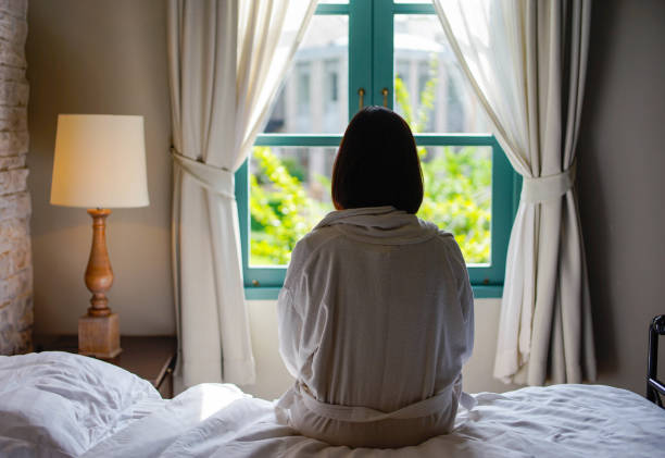 solitaria mujer sentada en la cama mirando a la ventana en la mañana - garment emotional stress equipment household equipment fotografías e imágenes de stock