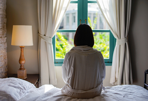 Lone woman sitting on the bed looking out at the window in the morning