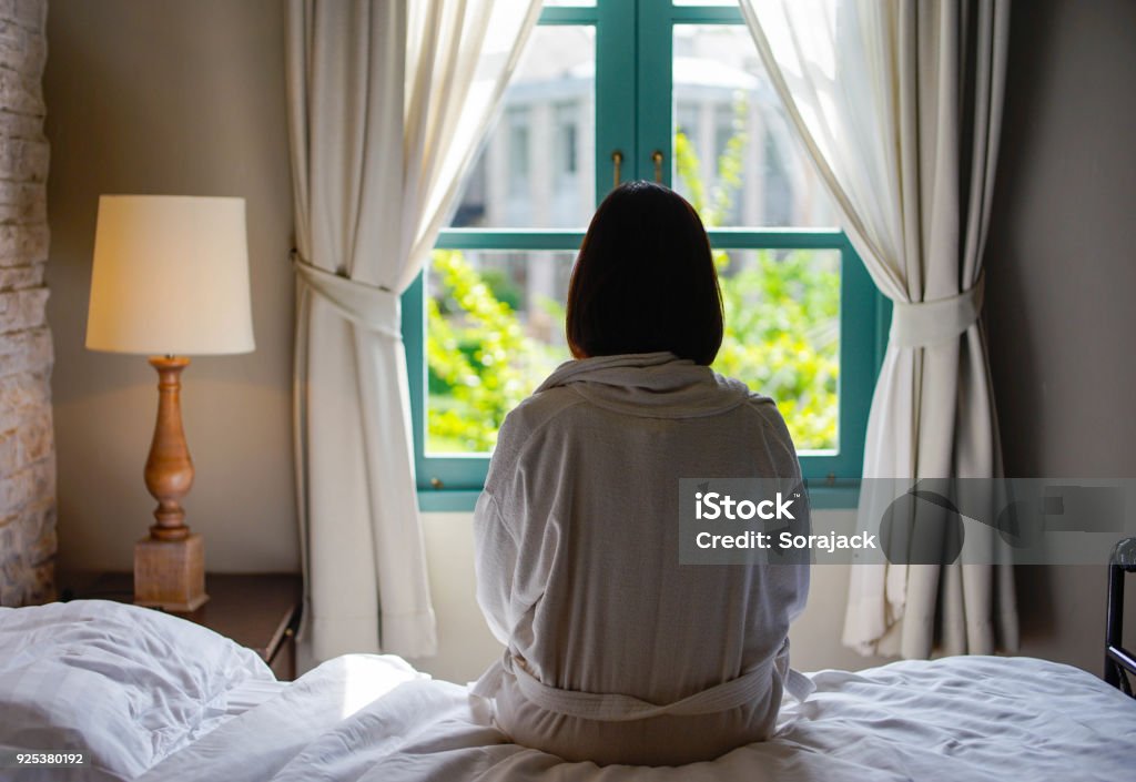Lone woman sitting on the bed looking out at the window in the morning Loneliness Stock Photo