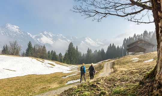 Two female friends enjoying a hike in early springtime with snow covered mountains in background. Reaching a mountain hut for a relaxing rest. Bavaria, Germany, Alps