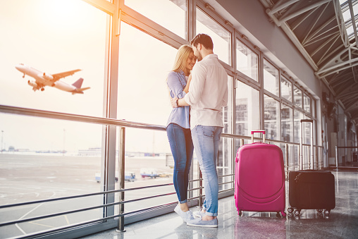 Romantic couple in airport. Attractive young woman and handsome man with suitcases are ready for traveling. Standing near panoramic windows while waiting for departure.