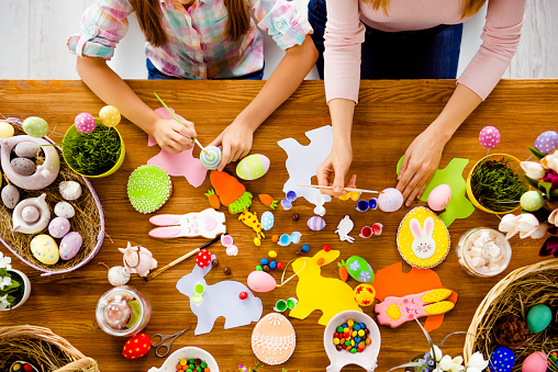 Top view of handmade easter objects, subjects, things, laying on wooden table, mother and daughter's hands painting, decorate eggs, holding brushed