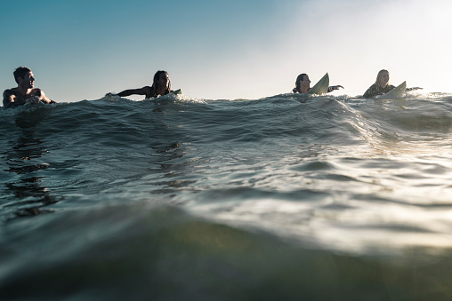 Portrait of a man with his surfboard standing in the sea making the shaka sign. He's looking at camera. Sunset time.