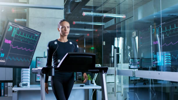 Beautiful Woman Athlete with Electrodes Connected to Her Body Walks on a Treadmill in a Sports Science Laboratory. In the Background High-Tech Laboratory with Monitors Showing EKG Readings. Beautiful Woman Athlete with Electrodes Connected to Her Body Walks on a Treadmill in a Sports Science Laboratory. In the Background High-Tech Laboratory with Monitors Showing EKG Readings. biomechanics stock pictures, royalty-free photos & images