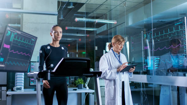 istock Woman Athlete Walks on a Treadmill while Scientist Holding Tablet Computer Supervises whole Process. In the Background High-Tech Laboratory with Analyzing Equipment and Monitors Showing EKG Readings. 925425546