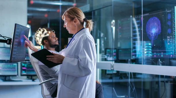 istock Man Wearing Brainwave Scanning Headset Sits in a Chair while Scientist Uses Touchscreen Display. In the Modern Brain Study Laboratory Monitors Show EEG Reading and Brain Model. 925425604
