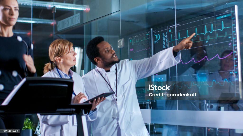 Beautiful Woman Athlete Runs on a Treadmill with Electrodes Attached to Her Body, while Two Scientists Supervise Watching EKG Data Showing on Laboratory Monitors. Hospital Stock Photo