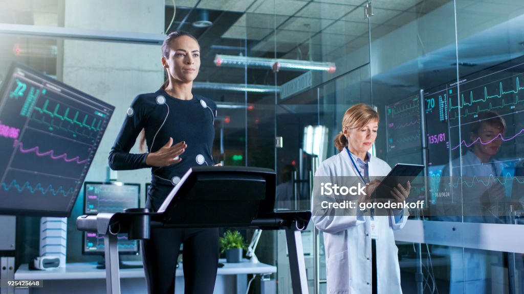 Woman Athlete Walks on a Treadmill  with Electrodes Attached to Her Body while Scientist Holding Tablet Computer Supervises whole Process. In the Background Laboratory with Monitors Showing EKG Readings. Athlete Stock Photo