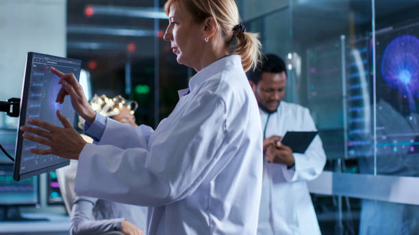 istock Woman Wearing Brainwave Scanning Headset Sits in a Chair while Scientist Uses Touchscreen Display, talks with Colleague. In the Modern Brain Study Laboratory Monitors Show EEG Reading and Brain Model. 925425652