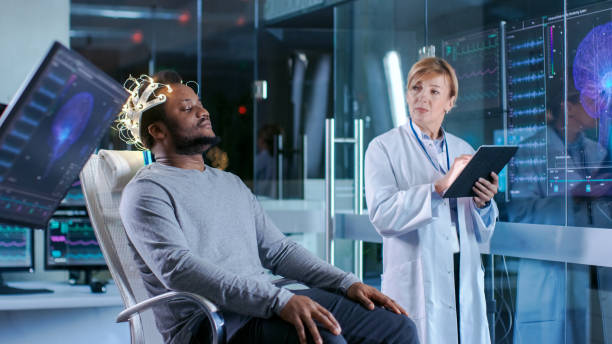 istock Man Wearing Brainwave Scanning Headset Sits in a Chair while Scientist with Tablet Computer Supervises Process. In the Modern Brain Study Laboratory Monitors Show EEG Reading and Brain Model. 925425662