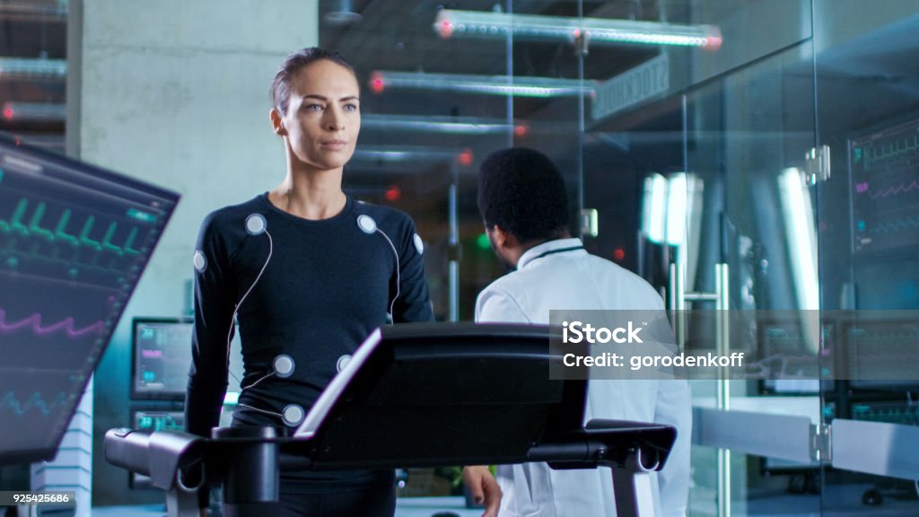 In Scientific Sports Laboratory Beautiful Woman Athlete Walks on a Treadmill with Electrodes Attached to Her Body, While Black Scientist Going Away, Monitors Show EKG Data on Display. Digital Tablet Stock Photo