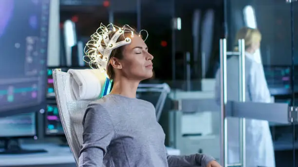 Woman Wearing Brainwave Scanning Headset Sits in a Chair while Scientist Supervises. In the Modern Brain Study Laboratory Monitors Show EEG Reading and Brain Model.