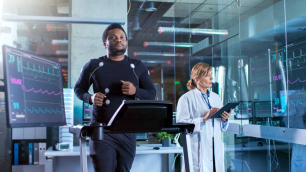 masculino o atleta caminha em uma esteira com eletrodos conectados ao seu corpo enquanto esporte cientista interage com touchscreen e supervisiona o status de ekg. no fundo laboratório com equipamentos de alta tecnologia. - biomecânica - fotografias e filmes do acervo