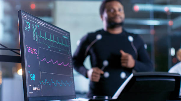 scatto ravvicinato di un monitor con dati ekg. l'atleta maschio corre su un tapis roulant con elettrodi attaccati al suo corpo mentre lo scienziato sportivo tiene il tablet e supervisiona lo stato dell'elettrocardiogramma in background. - electrode foto e immagini stock