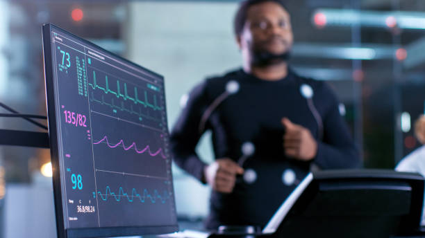 Close-up Shot of a Monitor With EKG Data. Male Athlete Runs on a Treadmill with Electrodes Attached to His Body while Sport Scientist Holds Tablet and Supervises EKG Status in the Background. Close-up Shot of a Monitor With EKG Data. Male Athlete Runs on a Treadmill with Electrodes Attached to His Body while Sport Scientist Holds Tablet and Supervises EKG Status in the Background. electrocardiography stock pictures, royalty-free photos & images