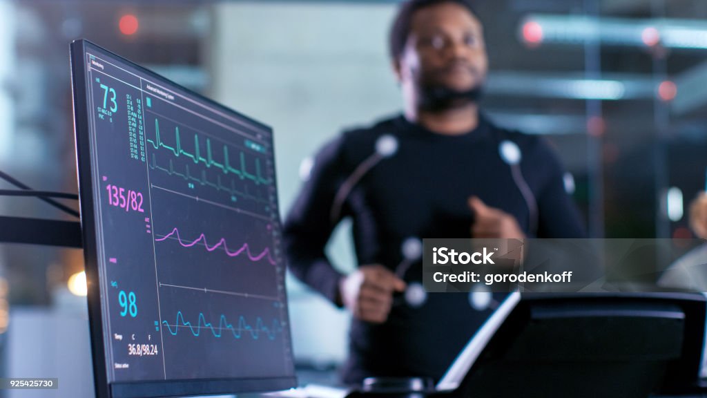 Foto de close-up de um Monitor com dados de ECG. Atleta masculino é executado em uma esteira com eletrodos conectados ao seu corpo enquanto esporte cientista mantém Tablet e supervisiona o Status de EKG no fundo. - Foto de stock de Ritmo Cardíaco royalty-free