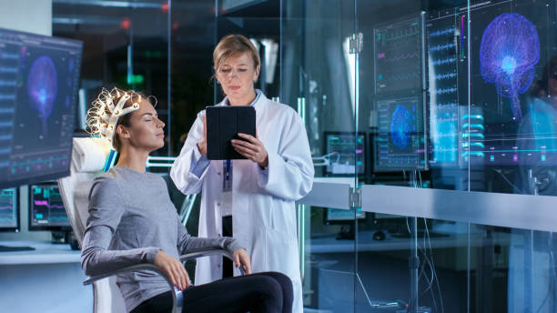 istock Woman Wearing Brainwave Scanning Headset Sits in a Chair while Scientist Adjusts the Device, Uses Tablet Computer. In the Modern Brain Study Laboratory Monitors Show EEG Reading and Brain Model. 925425738