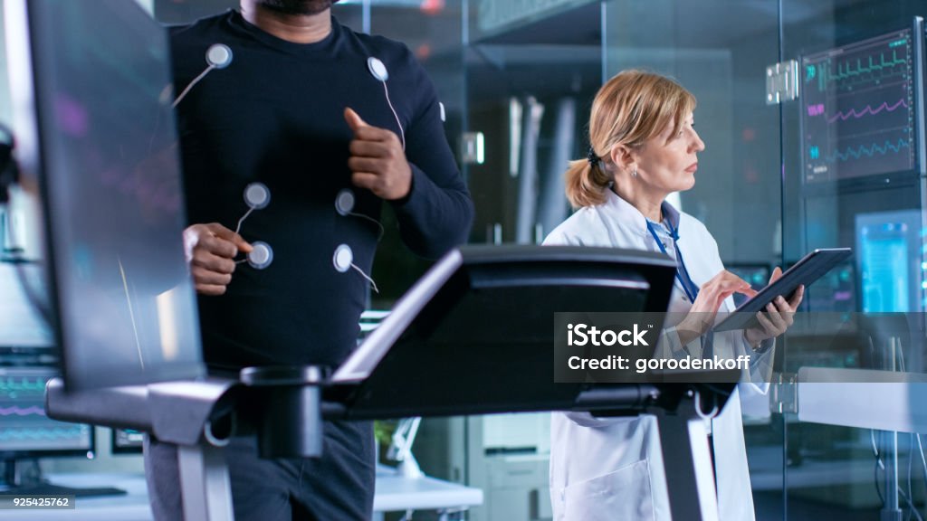 Monitor Shows EKG Reading of a Male Athlete Running on a Treadmill in the Background, Specialist Supervises Exercise Process, Controlling Physical Activity. Sport Science Theme. Laboratory Stock Photo