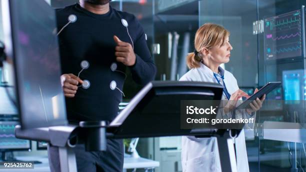 Monitor Shows Ekg Reading Of A Male Athlete Running On A Treadmill In The Background Specialist Supervises Exercise Process Controlling Physical Activity Sport Science Theme Stock Photo - Download Image Now
