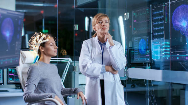 istock Woman Wearing Brainwave Scanning Headset Sits in a Chair while Scientist Adjusts the Device, Uses Tablet Computer. In the Modern Brain Study Laboratory Monitors Show EEG Reading and Brain Model. 925425798