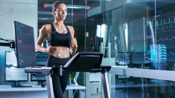 beautiful woman athlete runs on a treadmill with electrodes attached to her body, female physician uses tablet computer and controls ekg data showing on laboratory monitors. - physical checkup imagens e fotografias de stock