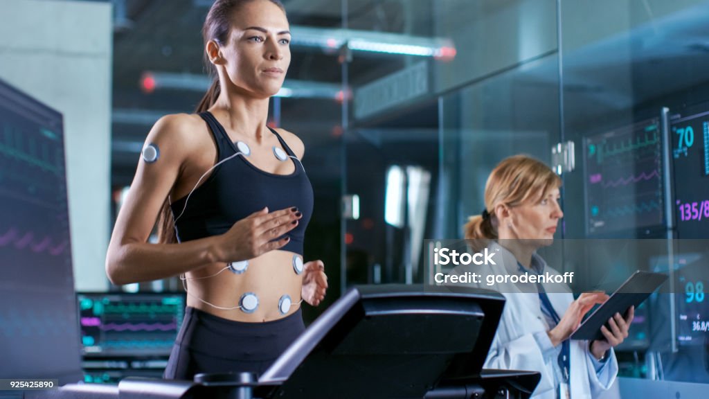 Beautiful Woman Athlete Runs on a Treadmill with Electrodes Attached to Her Body, Female Physician Uses Tablet Computer and Controls EKG Data Showing on Laboratory Monitors. Biomechanics Stock Photo